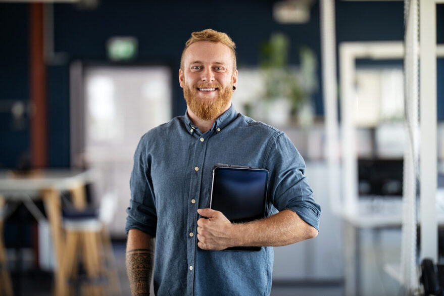 Ein junger Mann mit Bart und Tablet unter dem Arm in einem Büro