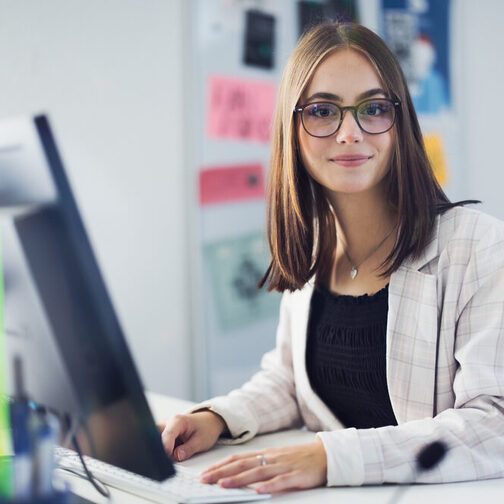 Eine junge Frau im Blazer sitzt in einem Büro vor einem PC und schaut freundlich in die Kamera. Im Hintergrund ist eine Pinnwand zu sehen.