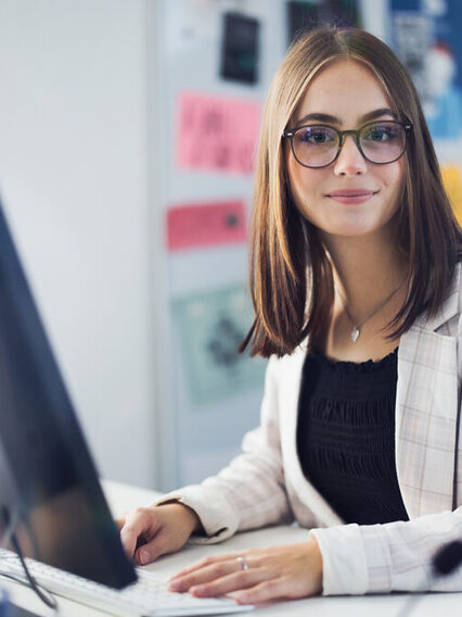 Eine junge Frau im Blazer sitzt in einem Büro vor einem PC und schaut freundlich in die Kamera. Im Hintergrund ist eine Pinnwand zu sehen.