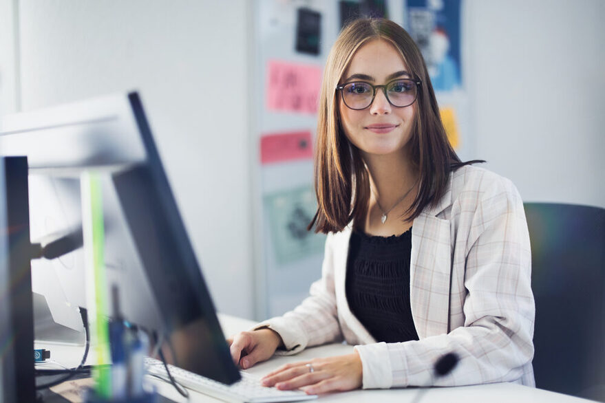 Eine junge Frau im Blazer sitzt in einem Büro vor einem PC und schaut freundlich in die Kamera. Im Hintergrund ist eine Pinnwand zu sehen.