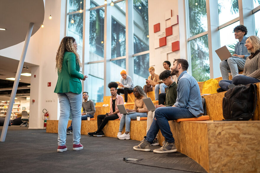 Ein Gruppe von Menschen sitzt in einer Bibliothek mit großen Fenstern im Hintergrund. Vor ihnen steht eine junge farbige Frau.