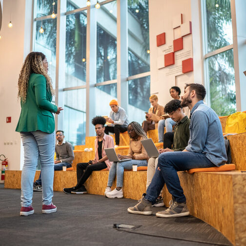 Ein Gruppe von Menschen sitzt in einer Bibliothek mit großen Fenstern im Hintergrund. Vor ihnen steht eine junge farbige Frau.