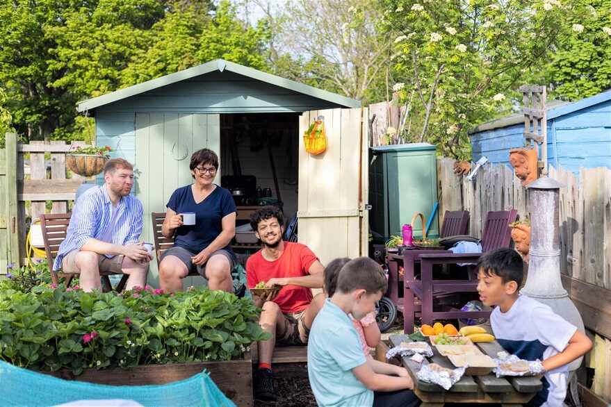 Drei Erwachsene verschiedenen Aussehens sitzen vor einem Schuppen machen eine Pause von der Gartenarbeit. Sie beobachten neugierig drei Kinder, die an einem Tisch sitzen, Obst essen und sich angeregt unterhalten..