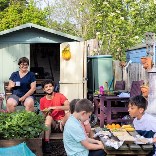 Drei Erwachsene verschiedenen Aussehens sitzen vor einem Schuppen machen eine Pause von der Gartenarbeit. Sie beobachten neugierig drei Kinder, die an einem Tisch sitzen, Obst essen und sich angeregt unterhalten..