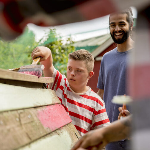 Ein Junge mit Down-Syndrom bemalt mit einem Pinsel Holz, ein junger Mann einer vermeintlich anderen ethnischen Herkunft steht hinter ihm und hilft ihm.