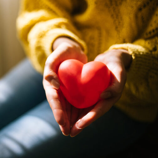 Die Hände einer sitzenden Frau, deren Gesicht auf dem Bildausschnitt nicht zu sehen ist, formen eine Mulde, in der ein rotes Herz liegt, was sie in die Kamera hält. Sie trägt eine Jeans und einen gelben Pullover mit Strickmuster.