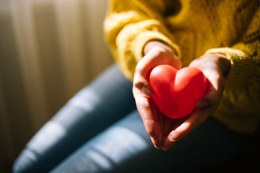 Die Hände einer sitzenden Frau, deren Gesicht auf dem Bildausschnitt nicht zu sehen ist, formen eine Mulde, in der ein rotes Herz liegt, was sie in die Kamera hält. Sie trägt eine Jeans und einen gelben Pullover mit Strickmuster.