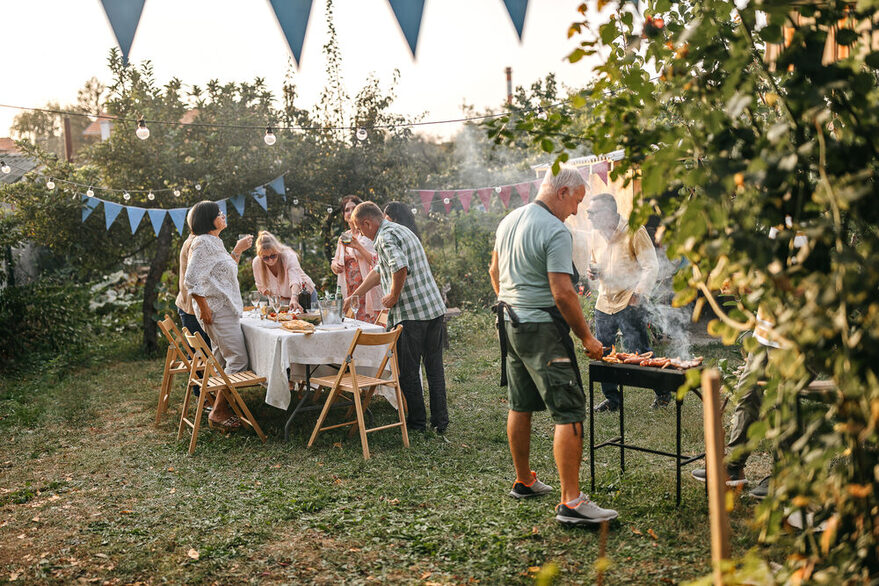 Gartenparty: Eine Gruppe bestehend aus sieben Menschen mittleren Alters trifft die letzten Vorbereitungen für eine Gartenparty im Sommer. Ein Mann auf der rechten Bildseite steht unter anderem mit Schürze bekleidet am Grill, von dem Rauch nach oben steigt. Der Großteil der Gruppe steht am Tisch, auf dem eine weiße Tischdecke liegt sowie verschiedene Speisen stehen und an dem Klappstühle aus Holz platziert sind (linke Bildseite). Der Garten ist mit Girlanden und Lichterketten dekoriert.