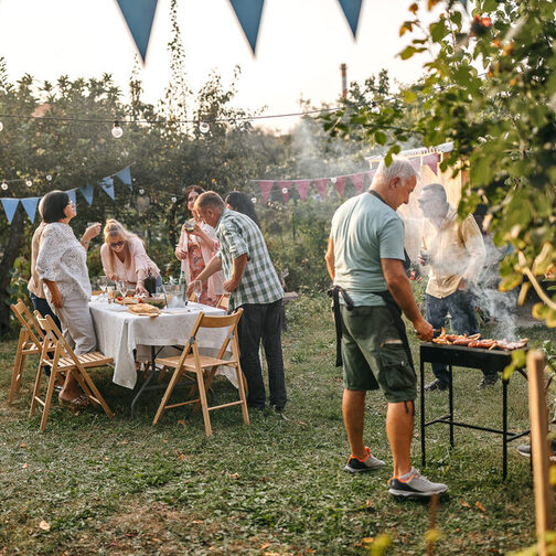 Gartenparty: Eine Gruppe bestehend aus sieben Menschen mittleren Alters trifft die letzten Vorbereitungen für eine Gartenparty im Sommer. Ein Mann auf der rechten Bildseite steht unter anderem mit Schürze bekleidet am Grill, von dem Rauch nach oben steigt. Der Großteil der Gruppe steht am Tisch, auf dem eine weiße Tischdecke liegt sowie verschiedene Speisen stehen und an dem Klappstühle aus Holz platziert sind (linke Bildseite). Der Garten ist mit Girlanden und Lichterketten dekoriert.