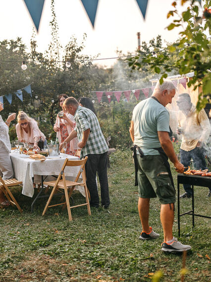 Gartenparty: Eine Gruppe bestehend aus sieben Menschen mittleren Alters trifft die letzten Vorbereitungen für eine Gartenparty im Sommer. Ein Mann auf der rechten Bildseite steht unter anderem mit Schürze bekleidet am Grill, von dem Rauch nach oben steigt. Der Großteil der Gruppe steht am Tisch, auf dem eine weiße Tischdecke liegt sowie verschiedene Speisen stehen und an dem Klappstühle aus Holz platziert sind (linke Bildseite). Der Garten ist mit Girlanden und Lichterketten dekoriert.