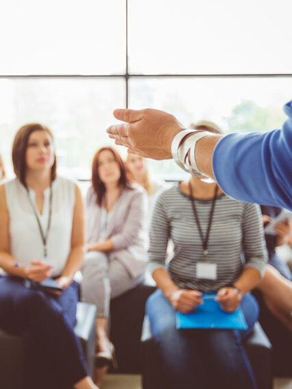 Workshop: Im Vordergrund auf der rechten Seite ist der linke Arm der Referentin zu sehen, die den Teilnehmerinnen zugewandt steht. Aufgeteilt in drei Reihen sitzen sie Teilnehmerinnen auf schwarzen Hockern und hören der Workshopleiterin aufmerksam zu.