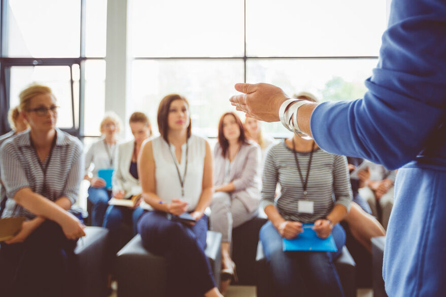 Workshop: Im Vordergrund auf der rechten Seite ist der linke Arm der Referentin zu sehen, die den Teilnehmerinnen zugewandt steht. Aufgeteilt in drei Reihen sitzen sie Teilnehmerinnen auf schwarzen Hockern und hören der Workshopleiterin aufmerksam zu.