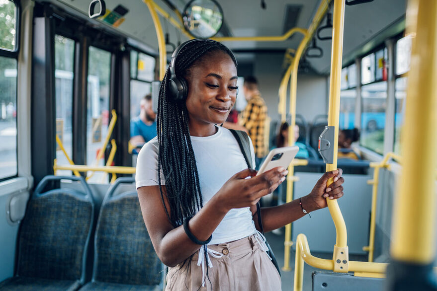 Eine Frau mit dunkler Haut steht im Bus und schaut mit fröhlichem Blick auf ihr Handy.