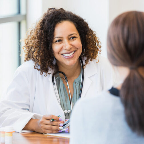 Eine freundlich lächelnde Ärztin mit dunklen, lockigen Haaren und einem Stethoskop um den Hals führt ein Gespräch mit einer Patientin. Sie sitzen sich an einem Tisch gegenüber. Die Ärztin ist von vorne, wohingegen die Patientin lediglich von hinten zu sehen ist.