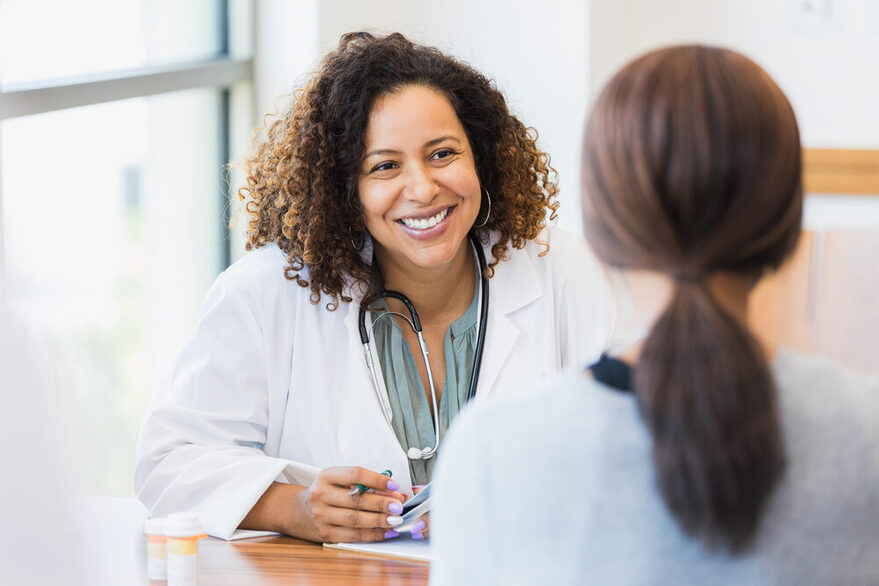 Eine freundlich lächelnde Ärztin mit dunklen, lockigen Haaren und einem Stethoskop um den Hals führt ein Gespräch mit einer Patientin. Sie sitzen sich an einem Tisch gegenüber. Die Ärztin ist von vorne, wohingegen die Patientin lediglich von hinten zu sehen ist.