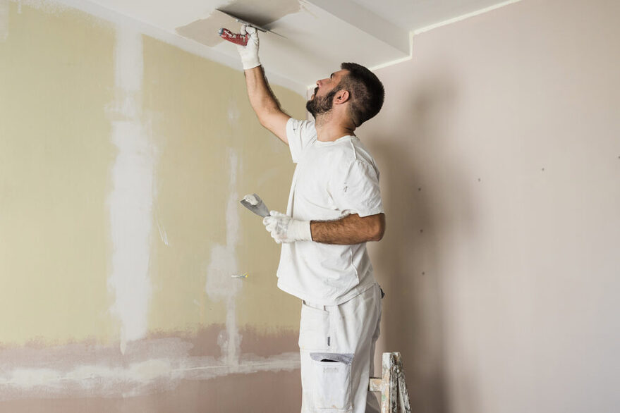 Mann mittleren Altes in Maler-Kleidung steht auf einer Leiter und spachtelt die Decke.