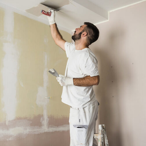 Mann mittleren Altes in Maler-Kleidung steht auf einer Leiter und spachtelt die Decke.