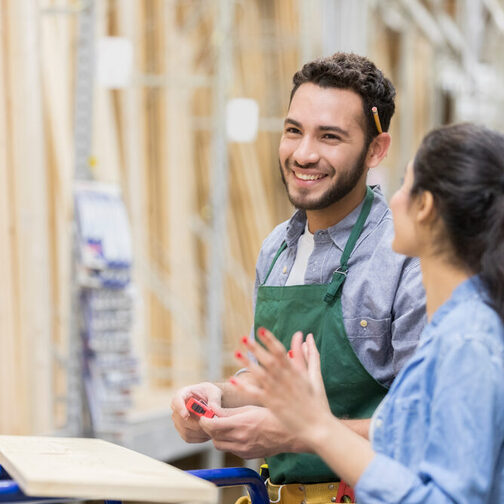 Ein junger Mann und eine junge Frau in Arbeitskleidung stehen gut gelaunt in einer Schreinerei. Vor ihnen liegt ein Holzbrett.