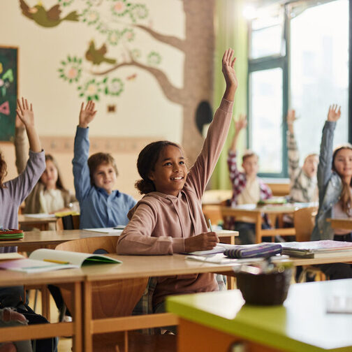 Fröhliche Grundschüler heben die Hände in einer Schulklasse.