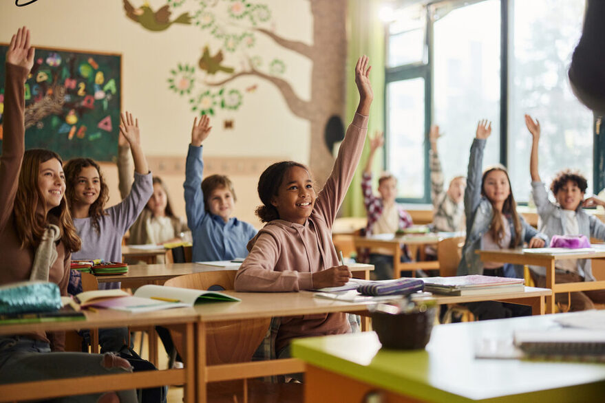 Fröhliche Grundschüler heben die Hände in einer Schulklasse.