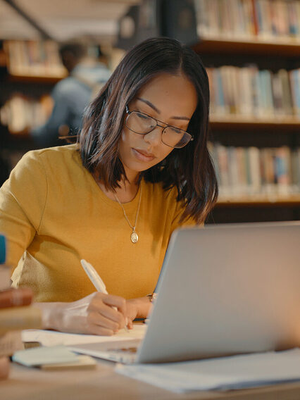 Junge Frau sitzt in einer Bibliothek an einem Schreibtisch. Vor ihr steht ihr Laptop drei Bücherstabel. Die Schreibtischlampe leuchtet.