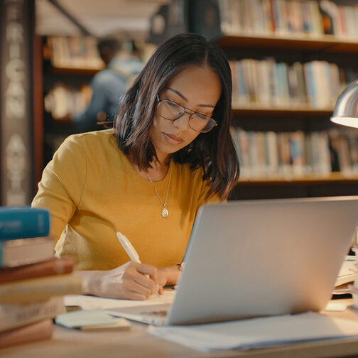 Junge Frau sitzt in einer Bibliothek an einem Schreibtisch. Vor ihr steht ihr Laptop drei Bücherstabel. Die Schreibtischlampe leuchtet.