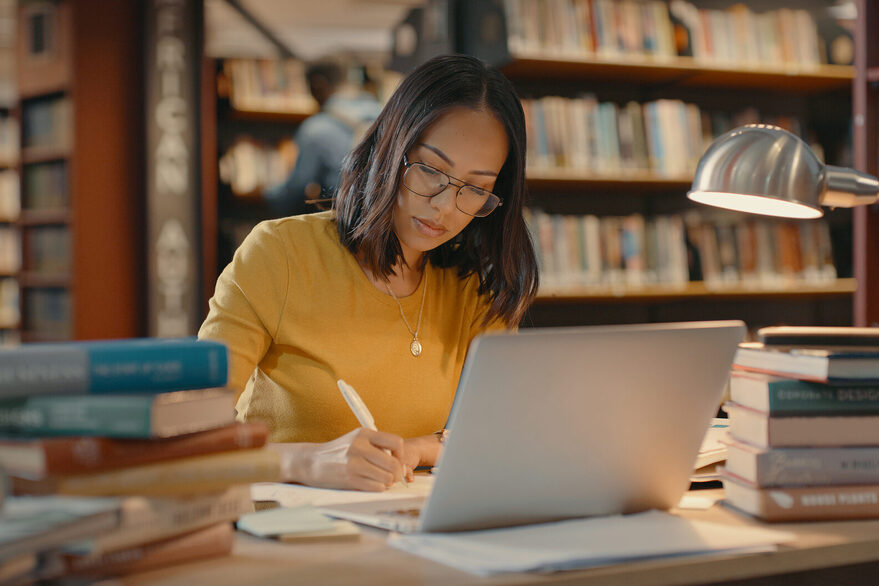 Junge Frau sitzt in einer Bibliothek an einem Schreibtisch. Vor ihr steht ihr Laptop drei Bücherstabel. Die Schreibtischlampe leuchtet.