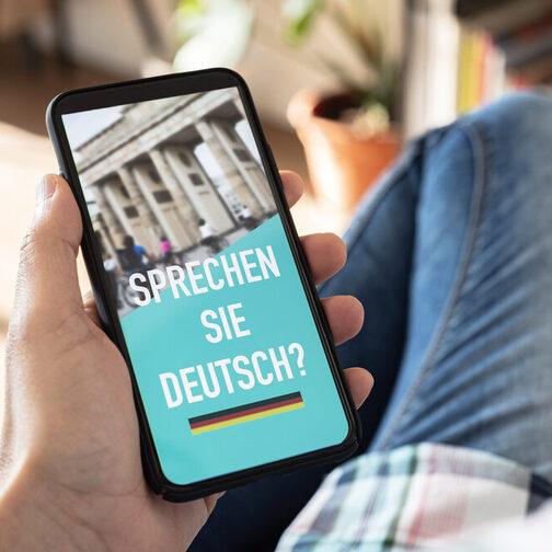 Ein Mann hält ein Handy in der Hand. Auf dem Bildschirm zu lesen ist die Frage "Sprechen Sie Deutsch?".