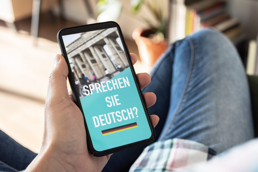 Ein Mann hält ein Handy in der Hand. Auf dem Bildschirm zu lesen ist die Frage "Sprechen Sie Deutsch?".