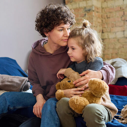 Eine junge Frau umarmt ihren kleinen Sohn, der einen braunen Teddybär in der Hand hat.