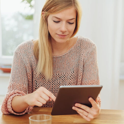 Eine junge Frau sitzt an einem Holztisch und tippt auf einem Tablet.