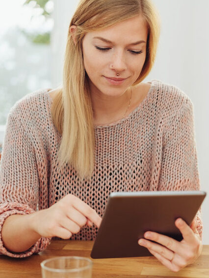 Eine junge Frau sitzt an einem Holztisch und tippt auf einem Tablet.