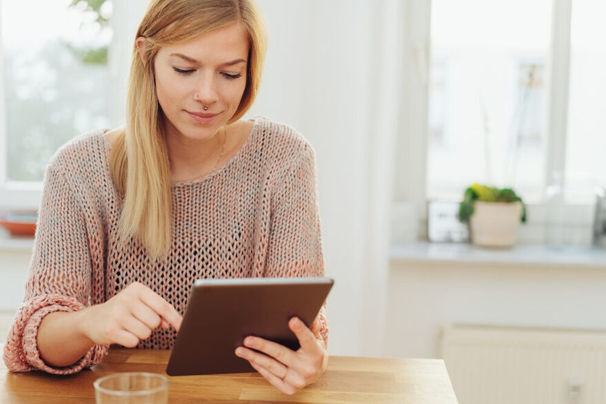 Eine junge Frau sitzt an einem Holztisch und tippt auf einem Tablet.